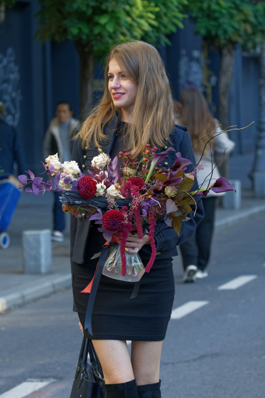 8 - A woman with long brown hair walking on a city street while carrying a large, rich autumn-toned floral bouquet and wearing a dark jacket, a black skirt, and knee-high boots